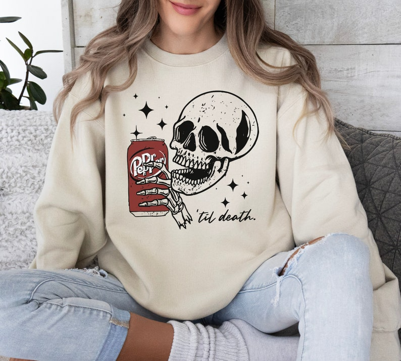 a woman sitting on a couch wearing a sweatshirt with a skull holding a can of